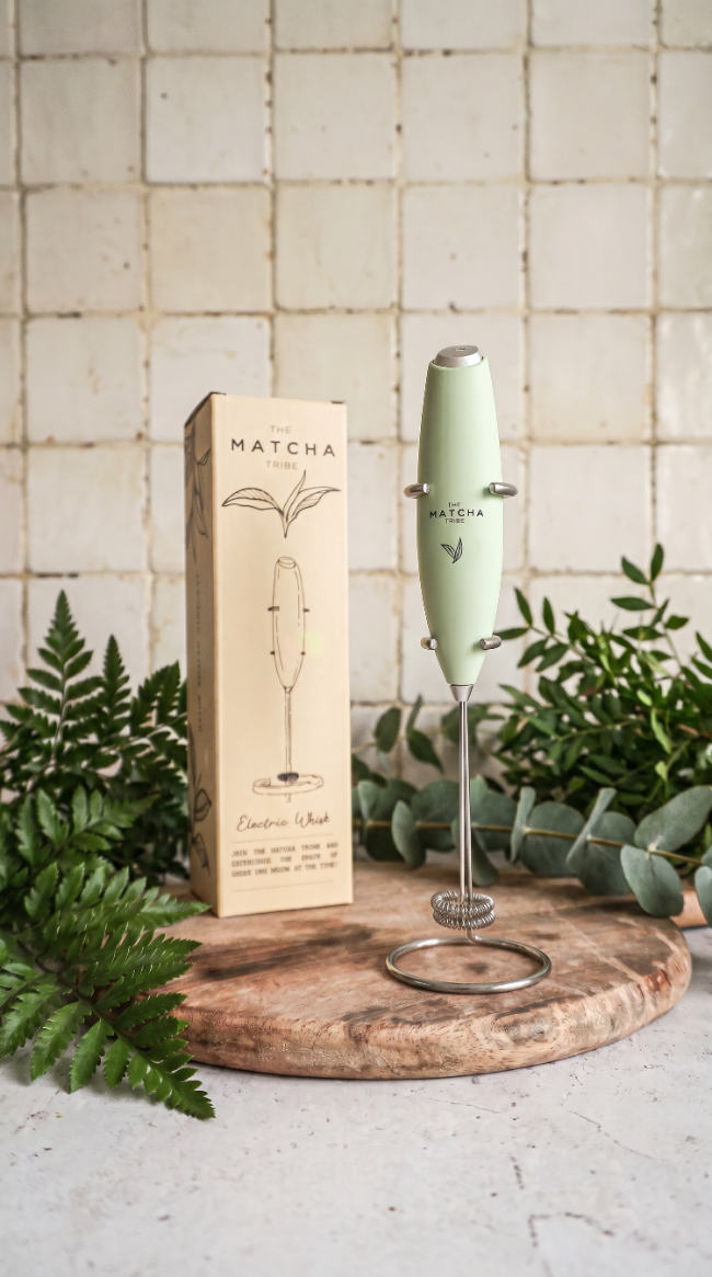 Matcha frother with packaging on a wooden board with plants in the background