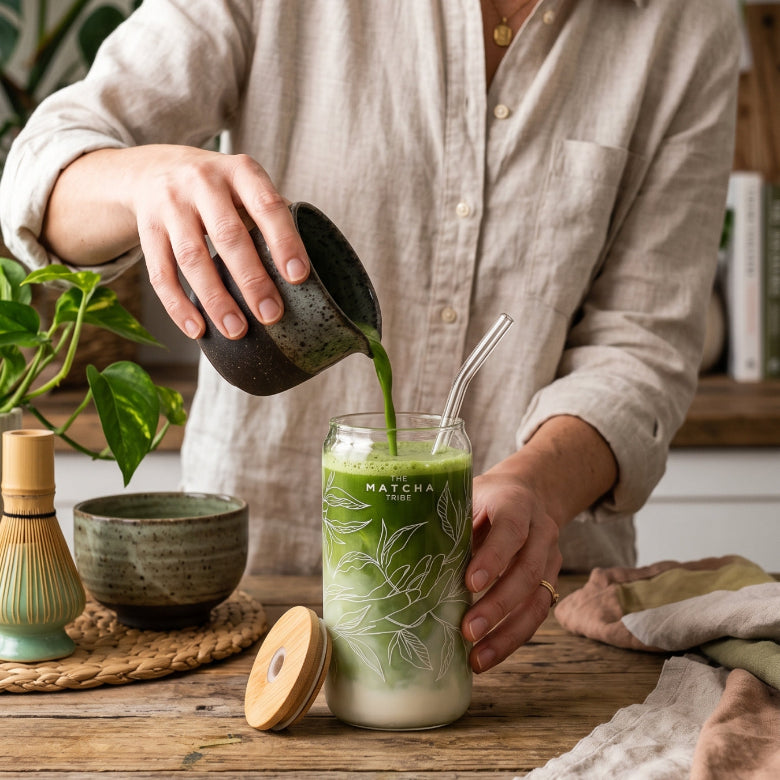 Person pouring green tea into a glass with a straw on a wooden table.