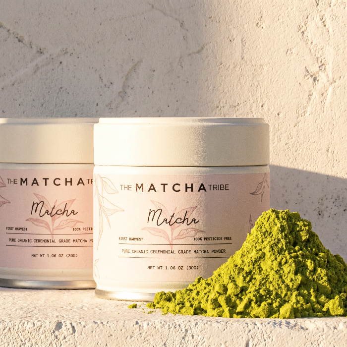 Two containers of The Matcha Tribe matcha powder with a pile of green matcha powder on a light background.